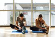 Eine Frau und ein Mann sitzen nebeneinander drinnen auf ihren Yogamatten auf dem Boden und dehnen beide ihr linkes ausgestrecktes Bein und ziehen den Oberen Teil des Schuhs mit dem linken Arm zu sich. Dabei ist das rechte Bein angewinkelt und die rechte Schuhunterseite berührt den inneren Oberschenkel.  Eine Frau und ein Mann sitzen nebeneinander drinnen auf ihren Yogamatten auf dem Boden und dehnen beide ihr linkes ausgestrecktes Bein und ziehen den Oberen Teil des Schuhs mit dem linken Arm zu sich. Dabei ist das rechte Bein angewinkelt und die rechte Schuhunterseite berührt den inneren Oberschenkel.